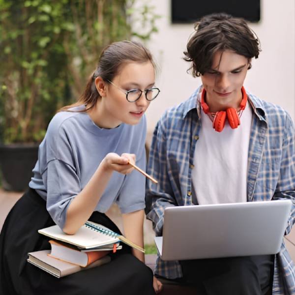 Photo of two students sitting on bench with books and working on laptop